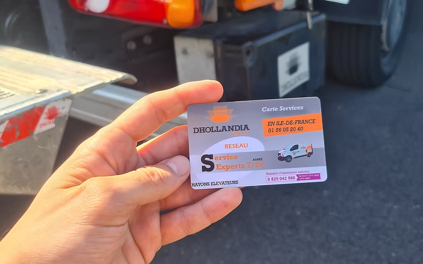 Carte Service en ligne Dhollandia Services tenue en main devant un hayon élévateur